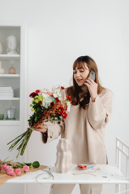 A young woman with long brown hair and bangs, wearing a beige sweater, is standing at a white table inside a bright, minimalist room. She is smiling and holding a large, colorful floral bouquet featur