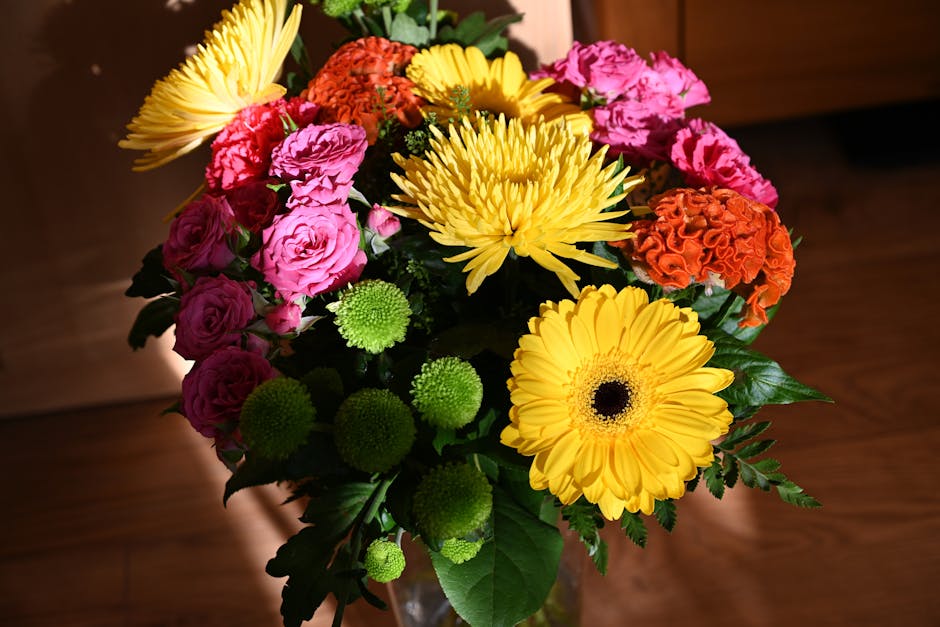 A vibrant floral arrangement in a clear glass vase featuring large yellow gerbera daisies with dark centers, bright pink roses with layered petals, orange carnations with ruffled blooms, and small gre